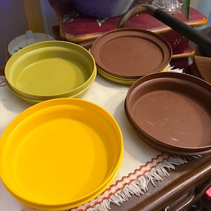 Può includere: Un set di tre contenitori rotondi in plastica con coperchio, composto da una ciotola giallo brillante, una ciotola verde oliva spento e una ciotola marrone scuro. I contenitori sono bassi con superfici lisce e coperchi circolari abbinati.