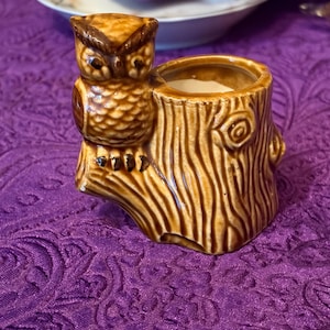 May include: A brown glazed ceramic planter shaped like a tree stump with a small owl perched on the side. The tree stump features textured bark details and a circular opening at the top, while the owl has detailed feathers and dark brown eyes.