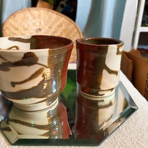 May include: Two handmade ceramic cups with a unique design. The cups feature a combination of brown, white, and green glaze. The cups are placed on a mirrored surface, reflecting their shapes. The cups are likely for drinking beverages.