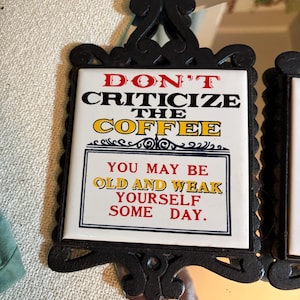 May include: A decorative sign with a black ornate frame and a mirrored base. The sign reads "DON'T CRITICIZE THE COFFEE" and "YOU MAY BE OLD AND WEAK YOURSELF SOME DAY" in red, yellow, and black text.