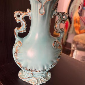 May include: A light blue ceramic vase featuring a flared, scalloped rim and two ornate, curved handles. The vase is decorated with gold-colored trim along the edges of the handles, the base, the rim, and the embossed, shell-like patterns.