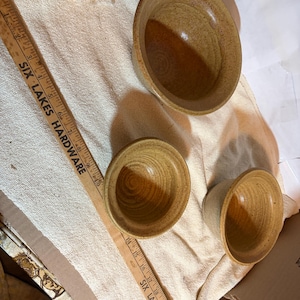 May include: Three handmade ceramic bowls in varying sizes, showcasing a warm, earthy color palette. The bowls are arranged on a textured, off-white fabric. A wooden ruler with the text "SIX LAKES HARDWARE" is visible.