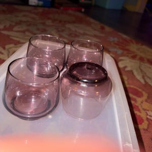 May include: A set of four small, rounded drinking glasses made of translucent purple glass. Three of the glasses are positioned upright, while one glass is placed upside down, revealing its circular base and smooth, curved rim design.