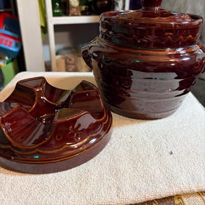 May include: A dark brown glazed ceramic bean pot with a matching lid and a separate circular base featuring a four-sectioned interior. The pot has a rounded body with a decorative embossed pattern around the rim and a small handle on the side.
