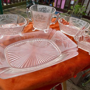 May include: A clear glass serving set with a rectangular tray and several bowls. The tray has the words "WE TRUST" etched into it. The set has a floral design and is sitting on an orange surface.