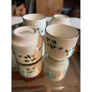 Op de afbeelding: Een set witte keramische eierdopjes met een groen klaverbladpatroon. De onderkant van een van de dopjes is duidelijk gemarkeerd met de tekst Carrigaline Pottery Co. Cork Ireland. Elk dopje heeft een goudkleurige rand en een glad, cilindrisch ontwerp.