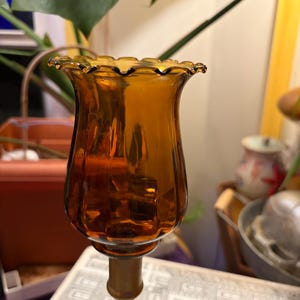Può includere: Un portacandele con un paralume in vetro ambrato traslucido con bordo arricciato e smerlato. Il paralume in vetro presenta pannelli scanalati verticali e poggia su una sottile base a piedistallo in metallo color ottone dal design arrotondato.