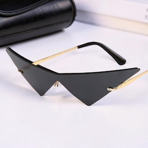 May include: Black geometric sunglasses with gold-colored arms and a black frame. The sunglasses have a unique triangular shape. A black case is visible in the background.