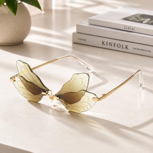 May include: Unique butterfly-shaped sunglasses with a gold metal frame. The lenses are a light amber color, and the butterfly wings have a delicate, veined design. The sunglasses are resting on a white surface.