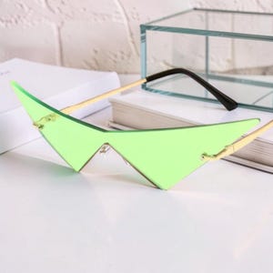 May include: Green-tinted sunglasses with a unique angular design. The lenses are a vibrant green, set in a gold-colored frame with black temple tips. The glasses are resting on a white surface.