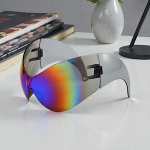 Op de afbeelding: Futuristische zilveren zonnebril met een regenboogkleurige lens. De zonnebril heeft een wrap-around design en een strakke, moderne uitstraling. De lenzen gaan over in een spectrum van kleuren, wat een levendig effect creëert. Het montuur is zilver van kleur.