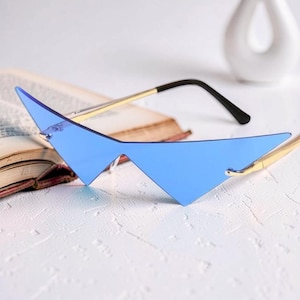 May include: Blue geometric sunglasses with gold-colored frames and black temples. The lenses have a sharp, angular design. The sunglasses are resting on a white surface with a book in the background.