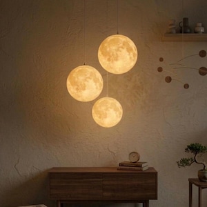 May include: Three spherical pendant lights resembling the moon hang from the ceiling. The lights are a warm yellow color, with detailed surface textures. The lights are suspended above a wooden cabinet, creating a cozy and inviting atmosphere.