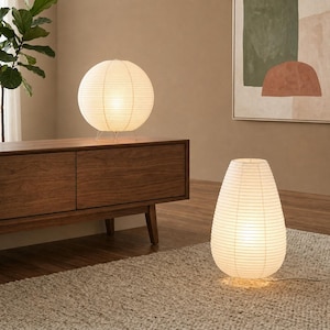 May include: Two paper lamps with a warm glow illuminate a room. One is spherical, mounted on a metal stand, and sits atop a wooden cabinet. The other is teardrop-shaped and rests on a beige rug. The lamps are made of white paper.