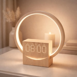 Könnte beinhalten: Ein digitaler Wecker aus Holz mit einem leuchtenden kreisförmigen Licht. Die Uhr zeigt "08:00" in weißen LED-Ziffern an. Das Design verfügt über einen gebogenen, beleuchteten Ring über dem Zifferblatt, der ein sanftes, umgebendes Leuchten erzeugt. Die Uhr steht auf einer weißen Oberfläche.