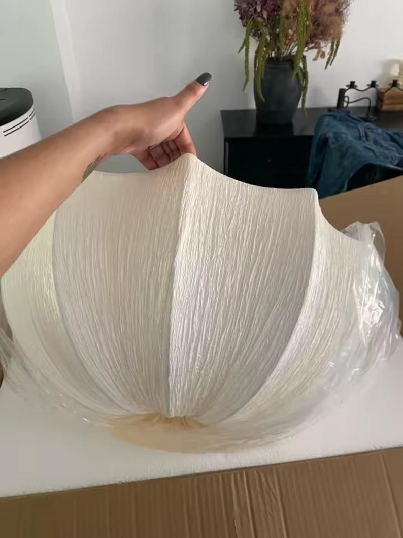 May include: A large, off-white, semi-circular lampshade with a distinctively crinkled, textured surface. The shade features a segmented, shell-like design with vertical ridges radiating from the center, partially encased in clear protective plastic.