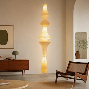 May include: A tall, vertical pendant light fixture with a stacked, multi-tiered silhouette made of a textured, off-white paper material. The lamp features several bulbous and tapered sections that emit a soft, warm glow from within the structure.