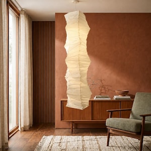 Pode incluir: Uma longa lanterna de papel branco sujo pende do teto numa sala de estar. A lanterna tem um design texturizado único e é iluminada por dentro. A sala apresenta um armário de madeira, uma poltrona verde e um tapete texturizado.