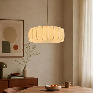 Puede incluir: Una lámpara colgante cilíndrica de color crema cuelga del techo. La luz tiene un diseño acanalado y emite un brillo suave y cálido. La lámpara está suspendida sobre una mesa de madera, creando un ambiente acogedor.