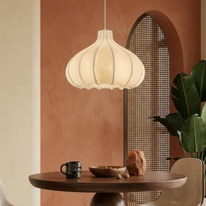 May include: Cream-colored pendant light with a unique, onion-shaped design, hanging above a round wooden table. The light has vertical seams and a scalloped bottom edge. A black mug, wooden bowl, and decorative stone are on the table.