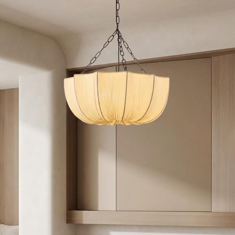 May include: A bowl-shaped pendant light fixture featuring a cream-colored fabric shade with vertical pleats and dark metal piping along the edges. The light is suspended from the ceiling by a dark metal chain that branches into three support arms.