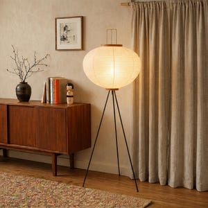 May include: A mid-century modern style floor lamp with a round, paper lantern shade. The lamp has a black tripod base and wooden accents. The lamp is lit, casting a warm glow. The lamp is in a room with a wooden cabinet and neutral-colored curtains.