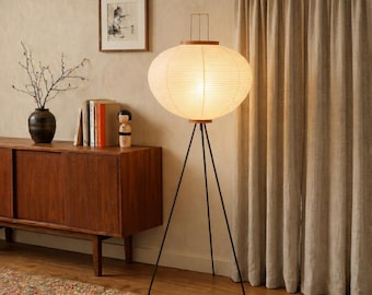 May include: A mid-century modern style floor lamp with a round, paper lantern shade. The lamp has a black tripod base and wooden accents. The lamp is lit, casting a warm glow. The lamp is in a room with a wooden cabinet and neutral-colored curtains.