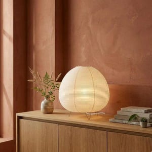 Peut inclure: Une lampe lanterne sphérique en papier crème dégageant une lueur chaleureuse, posée sur un meuble en bois. La lampe présente un design côtelé et est soutenue par de fins pieds argentés. Un petit vase avec de la verdure est à gauche.