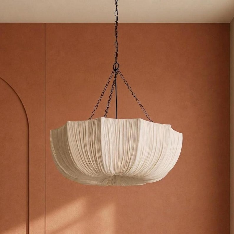 May include: A large, bowl-shaped pendant light fixture featuring a cream-colored fabric shade with vertical pleats. The shade is suspended by a thin black metal chain that connects to a central point, creating a structured, draped appearance.