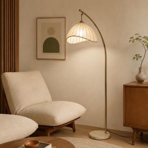 May include: A modern floor lamp with a curved, brass-colored metal frame and a cream-colored, pleated lampshade. The lamp is positioned in a living room setting, next to a cream-colored armchair and a wooden cabinet.