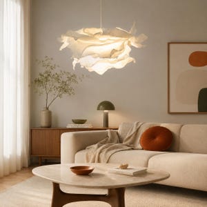 Puede incluir: Una lámpara colgante blanca con una pantalla en capas, con volantes y forma de nube suspendida del techo. La luz emite un brillo suave y cálido, iluminando los pliegues orgánicos y complejos del material que crean una apariencia voluminosa.