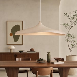 Puede incluir: Una lámpara colgante ancha de color blanco roto con una forma curva y lisa similar a un platillo, suspendida de un cable blanco fino. La luminaria presenta una superficie mate y texturizada que difunde la luz hacia abajo, creando un brillo suave.