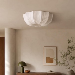 Peut inclure: Un plafonnier en tissu blanc plissé. La lumière a une forme arrondie et festonnée et est montée sur un plafond de couleur neutre. La pièce comprend une armoire en bois, une lampe verte et une œuvre d'art abstraite encadrée.