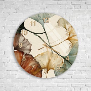 Può includere: Un orologio da parete rotondo con sfondo color crema decorato con grandi foglie di ginkgo sovrapposte nei toni del ruggine, oro, verde salvia e verde acqua. Il quadrante presenta grandi numeri chiari da 1 a 12 e lancette sottili.