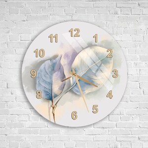 Puede incluir: Un reloj de pared redondo con esfera blanca que presenta una ilustración en acuarela de dos hojas en tonos azul y crema. El reloj tiene números dorados del 1 al 12 y finas manecillas doradas para las horas, los minutos y los segundos.