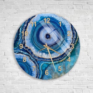 May include: A round wall clock featuring a blue agate stone pattern with concentric rings in shades of navy, azure, and white. The clock face displays gold-colored Arabic numerals from 1 to 12 and thin gold-colored hour, minute, and second hands.