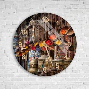 Könnte beinhalten: Eine runde Wanduhr mit einem Fotodruck eines Gewürzmarktes, der hängende Metallschalen mit bunten Gewürzen und Messingbehälter zeigt. Das Zifferblatt hat goldfarbene Zahlen von 1 bis 12 sowie dünne, goldfarbene Zeiger für die Uhrzeit.