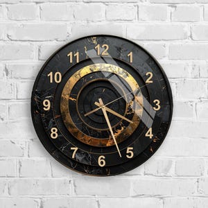 Puede incluir: Un reloj de pared redondo con esfera negra efecto mármol y detalles dorados. El reloj presenta manecillas y números dorados, con un diseño concéntrico. Está montado sobre una pared de ladrillo blanco.