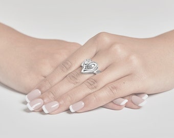 May include: A silver ring with a heart-shaped design featuring intricate swirls. The ring is worn on the middle finger of a hand with manicured nails.