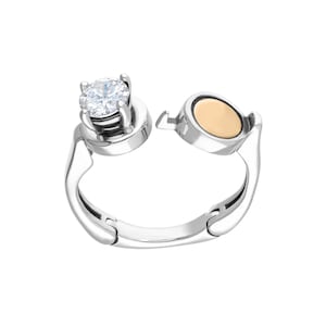 May include: A silver ring with a clear stone and a gold-toned accent. The ring is open and has a hinged design.