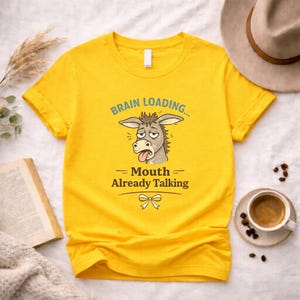 May include: Bright yellow t-shirt featuring a cartoon donkey with its tongue out, and the text "Brain Loading... Mouth Already Talking." The shirt has a crew neck and is a vibrant yellow colour.