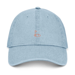 May include: Pale blue denim baseball cap with a curved peak and adjustable strap. The cap has an embroidered orange golf flag design. A casual accessory for golf lovers.