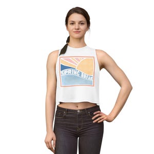 May include: White crop top with a colorful graphic that says "SPRING BREAK". The graphic features a sun, ocean, and sky in shades of orange, pink, blue, and peach. The top is sleeveless and has a round neckline.