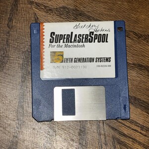 May include: A blue 3.5-inch floppy disk with a white label featuring the text SuperLaserSpool For the Macintosh and Fifth Generation Systems. The label includes a handwritten name, a serial number 912-0021130, and a code 330-01216-300.