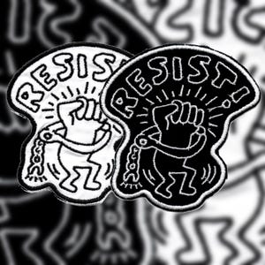 May include: Two embroidered patches with the word "RESIST" and a stylized figure with a raised fist. One patch is black and white, the other is white with black outlines. The design features a chain and radiating lines.