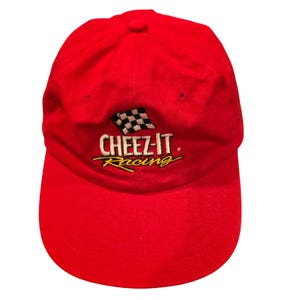 May include: A bright red baseball cap featuring an embroidered logo on the front. The logo includes a black and white checkered flag above the white text CHEEZ-IT and the yellow cursive word Racing, all centered on the crown of the hat.