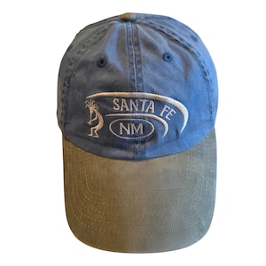 Puede incluir: Una gorra de béisbol azul con visera color canela. La parte frontal presenta un bordado blanco de una figura Kokopelli, el texto SANTA FE y un óvalo con las letras NM, enmarcado por un diseño de línea blanca curva.