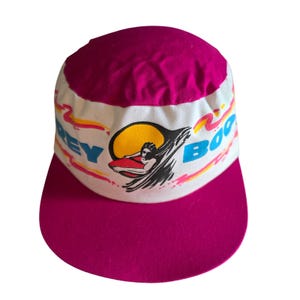 Puede incluir: Una gorra de béisbol magenta y blanca con un gráfico de una persona surfeando sobre una ola frente a un sol amarillo. El texto "GREY BOO" está impreso en azul en el panel frontal blanco, acentuado por líneas onduladas amarillas y rosas.