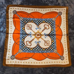 May include: A square scarf featuring a central cream-colored panel with four ornate gold keys arranged in a cross pattern, accented by light blue bows. The design includes a blue and white checkered border, orange sections with chain motifs, and a brown edge.