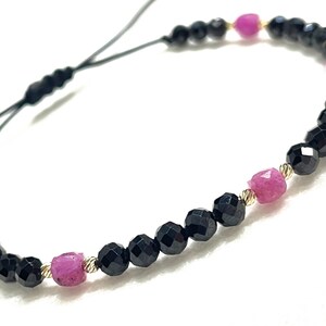 May include: A delicate bracelet featuring alternating black faceted beads and vibrant pink gemstones. The bracelet is strung on a black cord and includes gold-colored accents, creating a striking contrast of colors and textures. The bracelet is designed to be adjustable.